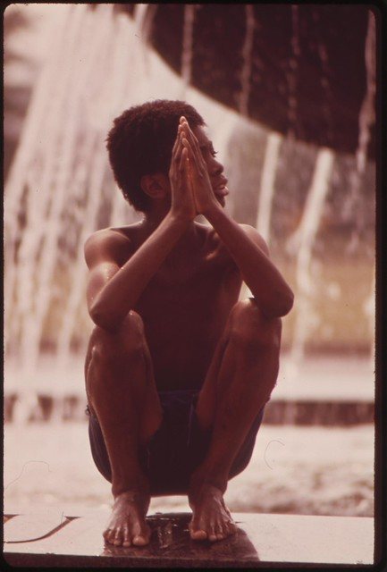 A boy crouching at the edge of a fountain, hands raised, North Philadelphia, 1973