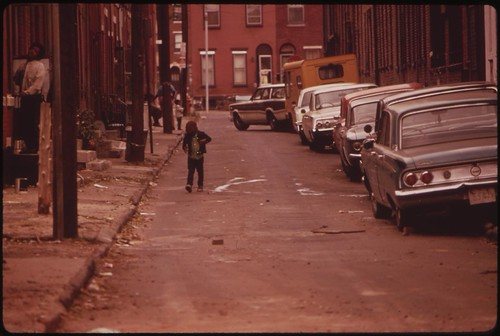 A small child walking down a North Philadelphia block lined with rowhouses and parked cars, 1973