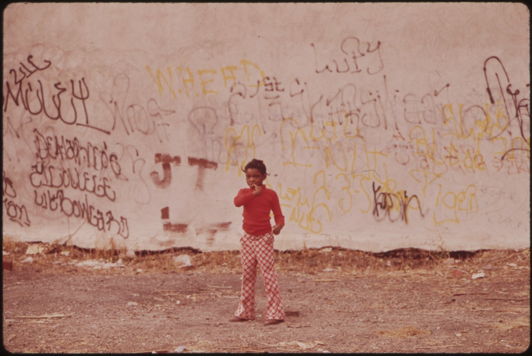 A young child in a red shirt and red plaid pants standing before a graffiti-covered wall, North Philadelphia, 1973