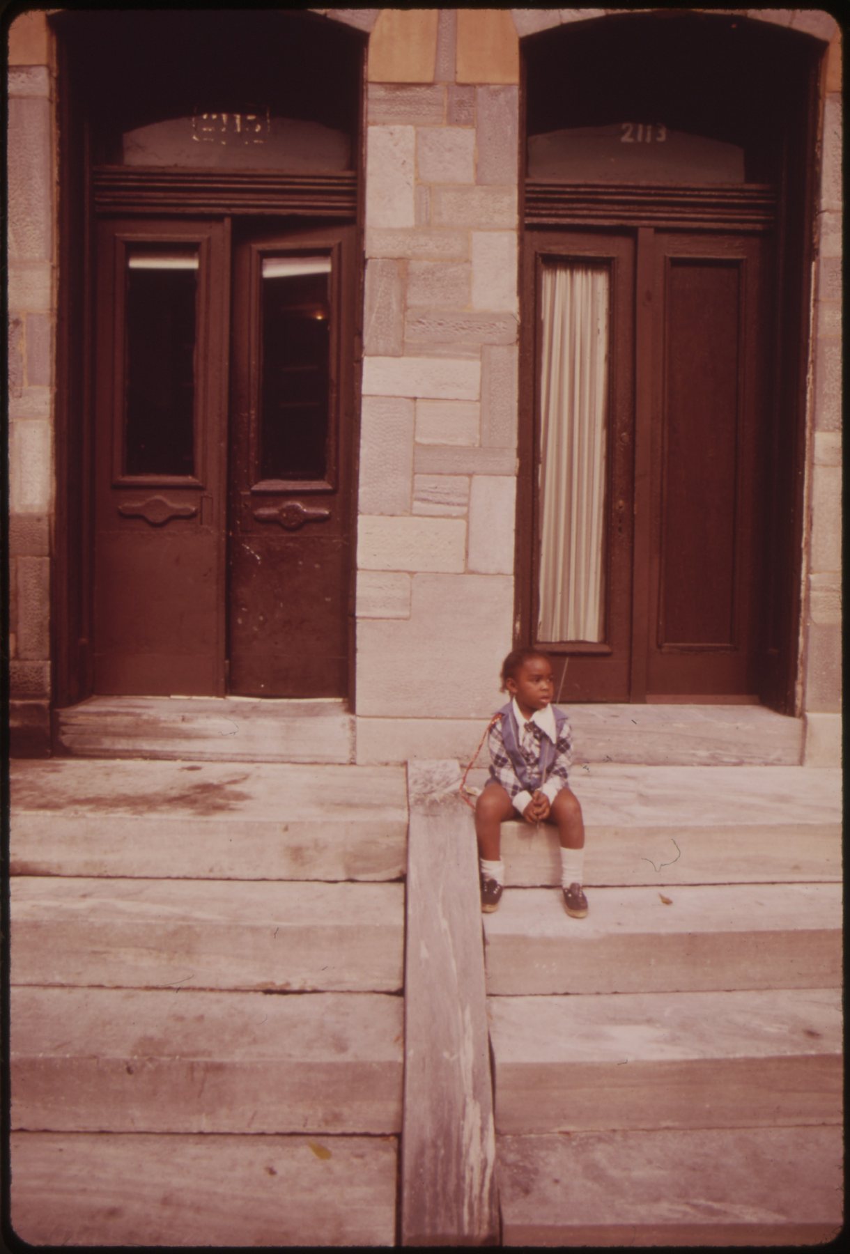 A young girl in an argyle vest sitting on the stone steps of a North Philadelphia rowhouse, 1973