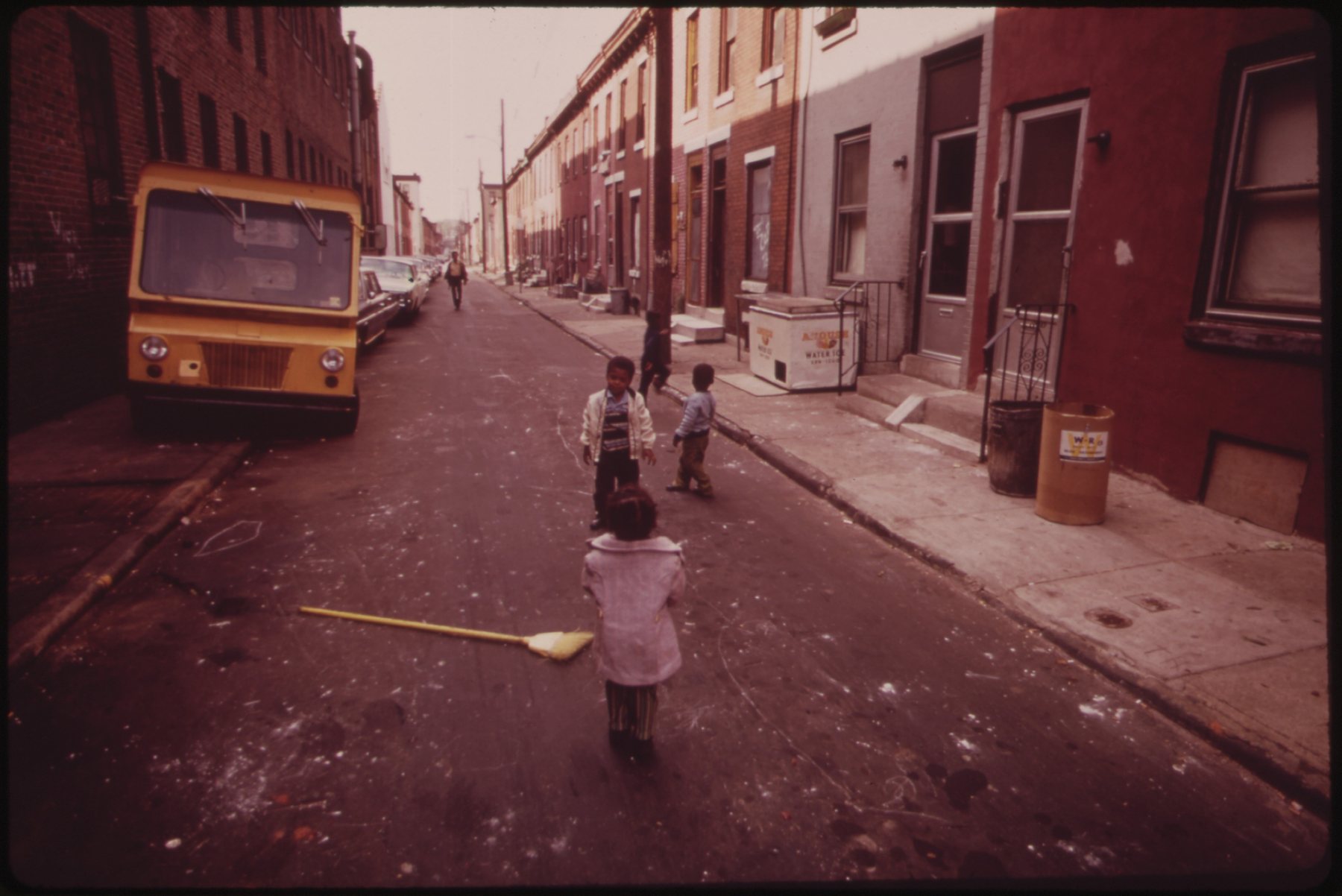 Kids playing in the street beside a yellow van and red brick rowhouses, North Philadelphia, 1973