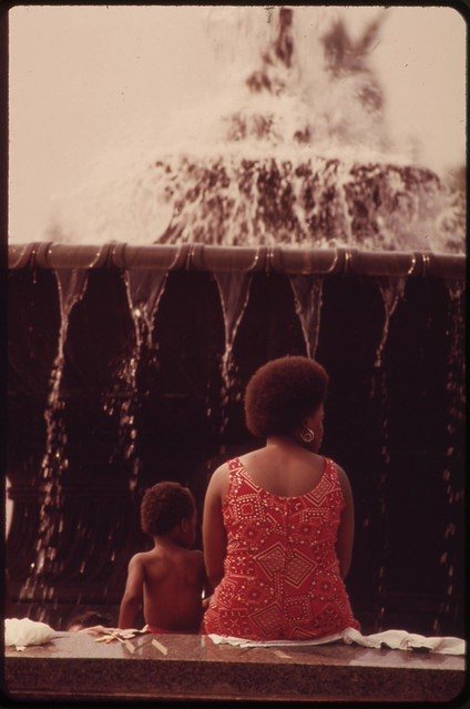 A mother in a patterned dress and her young son at the edge of a fountain, backs to the camera, North Philadelphia, 1973