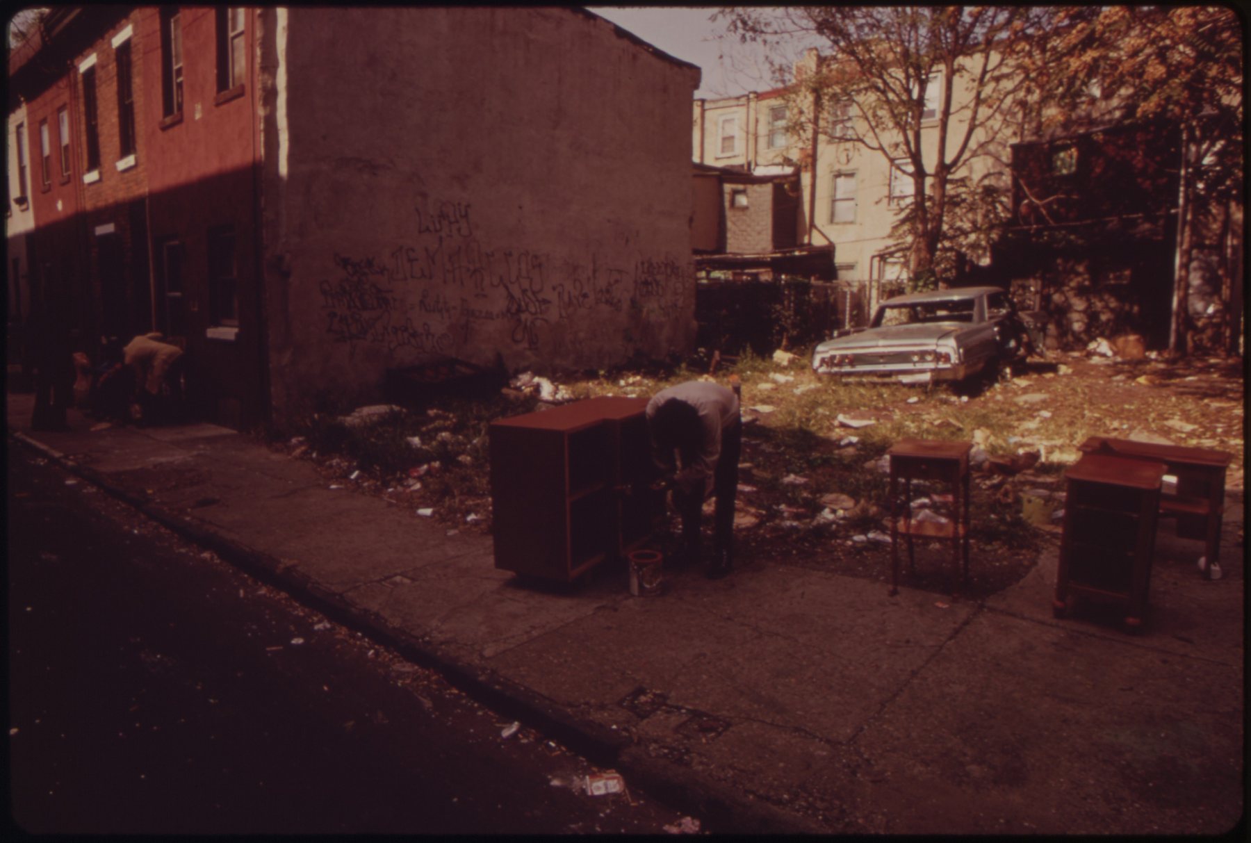 A man in a vacant lot with an abandoned car and salvaged furniture, North Philadelphia, 1973